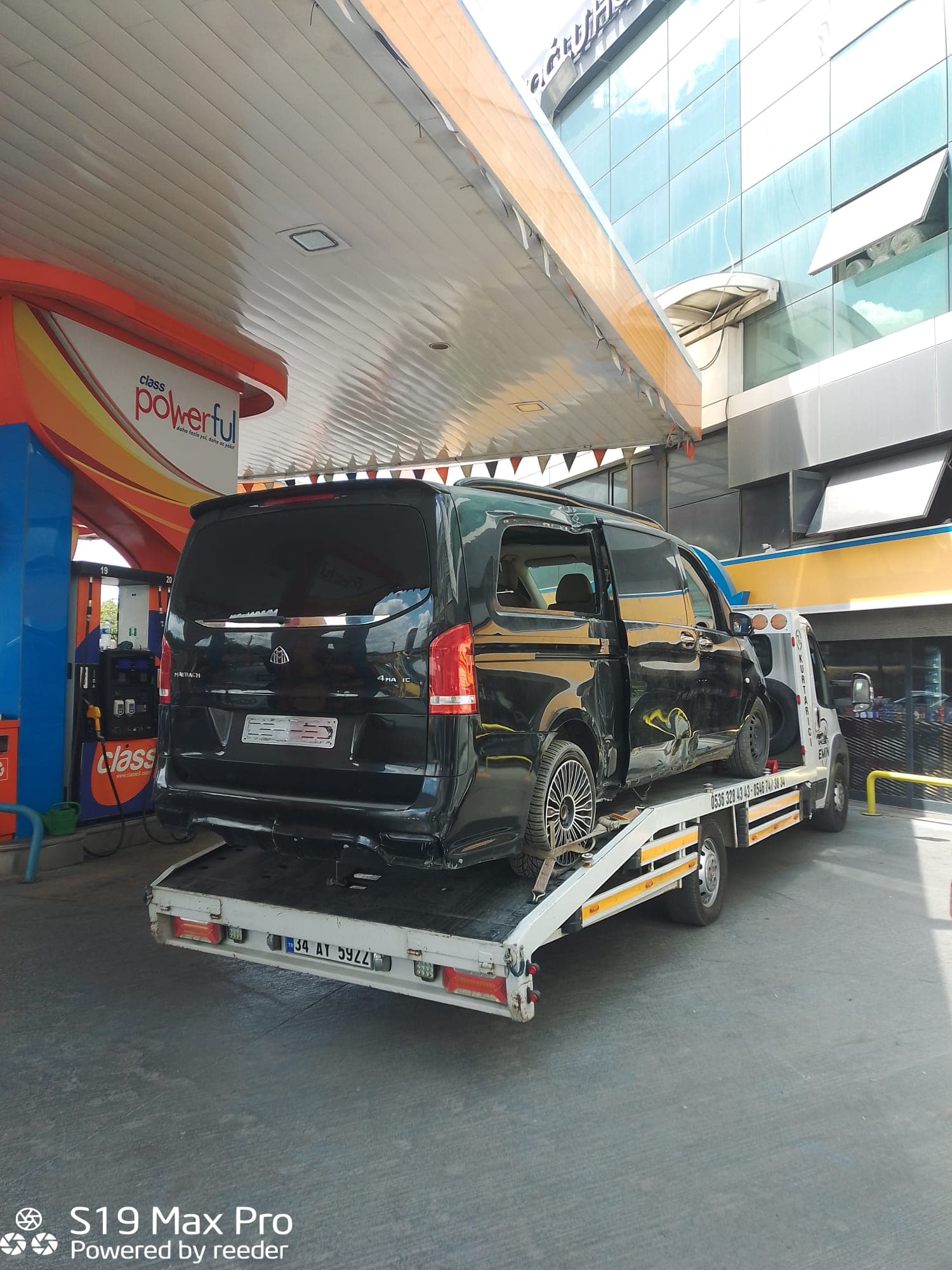 Yan tarafı ağır hasar görmüş siyah minivan, çekici platformuna yüklenmiş halde akaryakıt istasyonunda